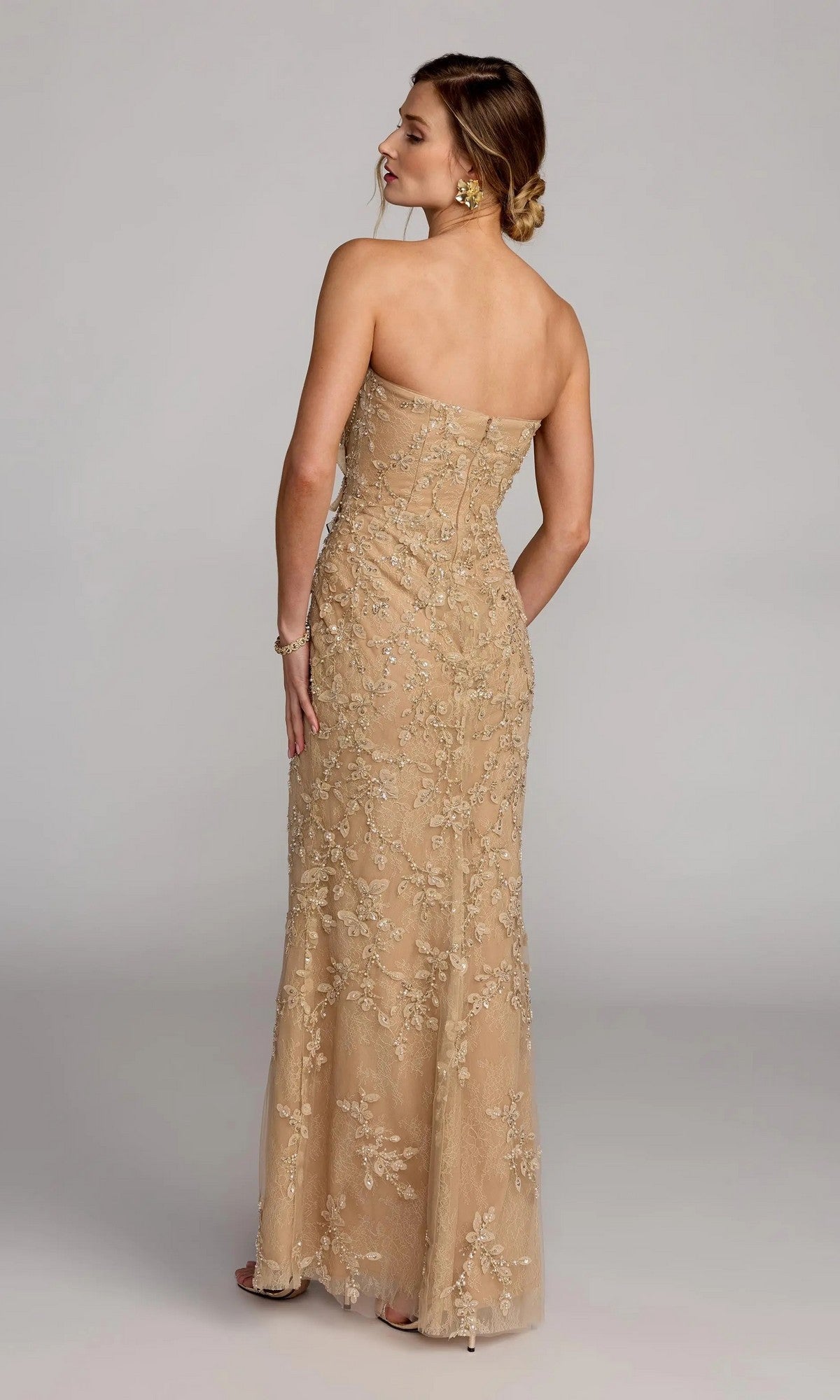 A woman faces away, wearing the Mac Duggal 21200 long formal dress—a gold strapless gown with floral embroidery. Her hair is in a low bun, styled with earrings and a bracelet for an elegant look.